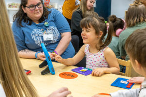 A young girl is smiling and using a speech generating device in a classroom.